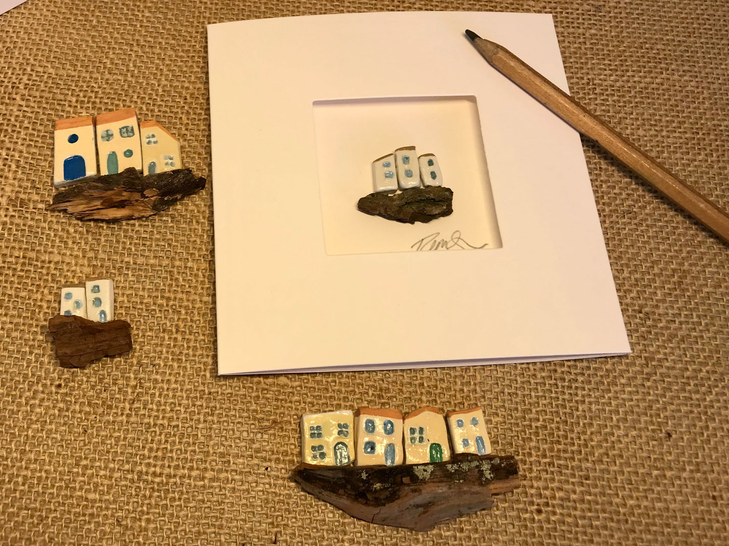 Customizable 'Little Worlds' card featuring a single handcrafted clay house on driftwood, surrounded by various miniature house styles to showcase customization options; a pencil included for scale.