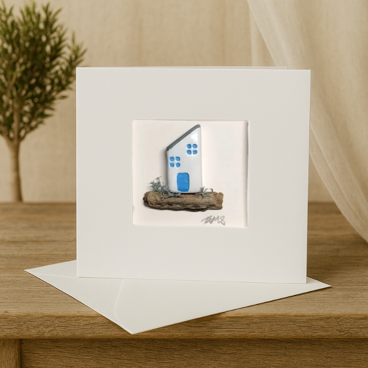 Handmade coastal cottage greeting card featuring a white house with blue details, set on driftwood, displayed with matching envelope.