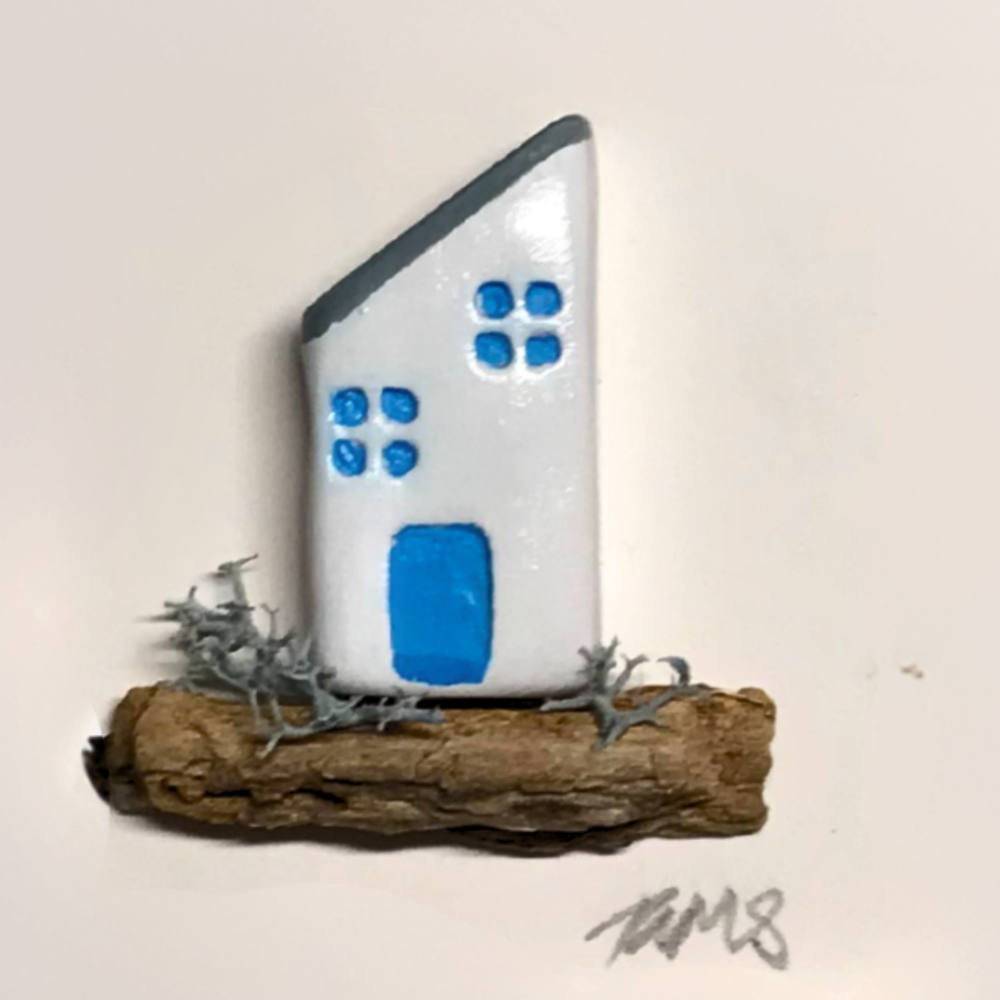Close-up of Pebble Cottage design, featuring a white house with sloping roof, blue windows and door, set on a natural driftwood base
