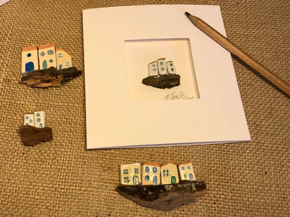 Customizable 'Little Worlds' card featuring a single handcrafted clay house on driftwood, surrounded by various miniature house styles to showcase customization options; a pencil included for scale.
