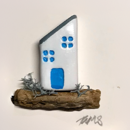 Close-up of Pebble Cottage design, featuring a white house with sloping roof, blue windows and door, set on a natural driftwood base